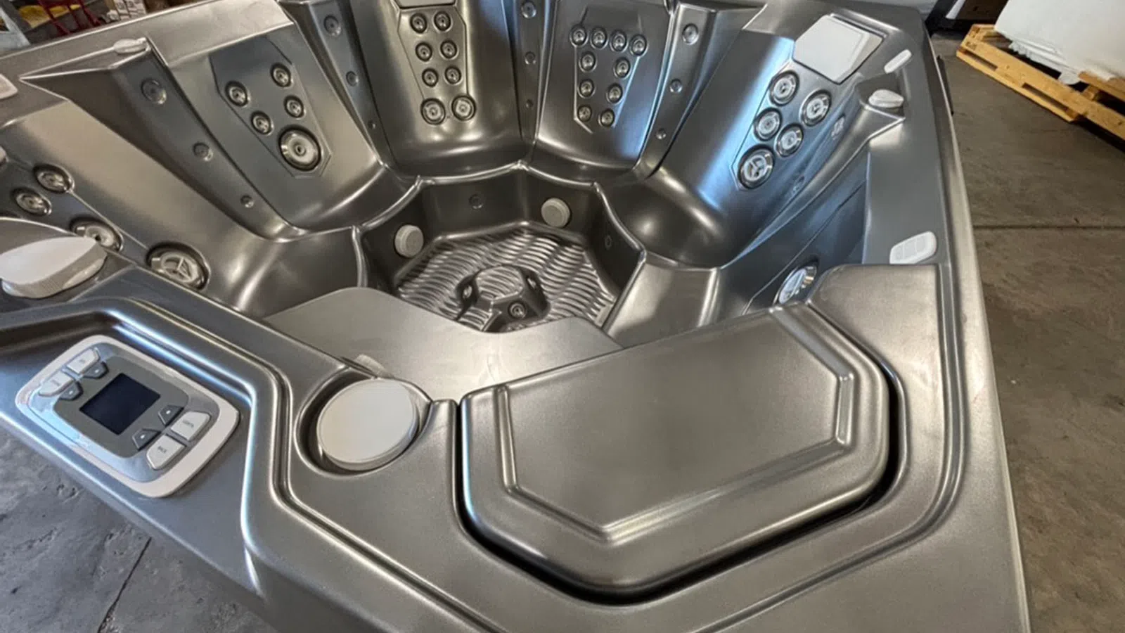 Silver multi-seat hot tub with numerous jets and control panel in an indoor warehouse setting.