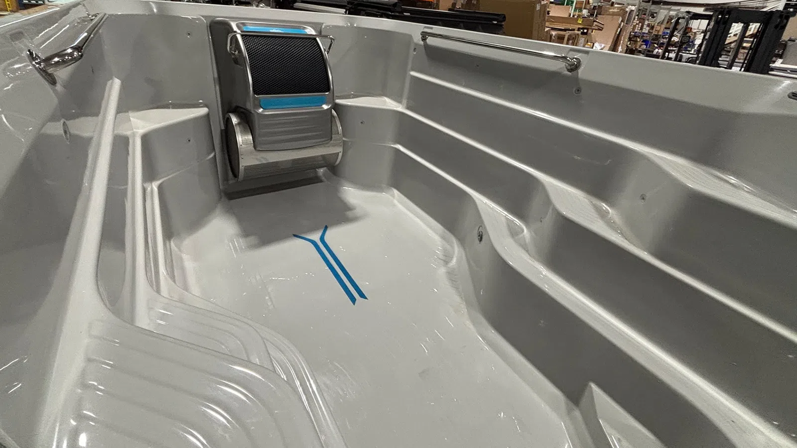 Empty gray fiberglass hot tub shell with built-in seating and a headrest inside a warehouse setting.