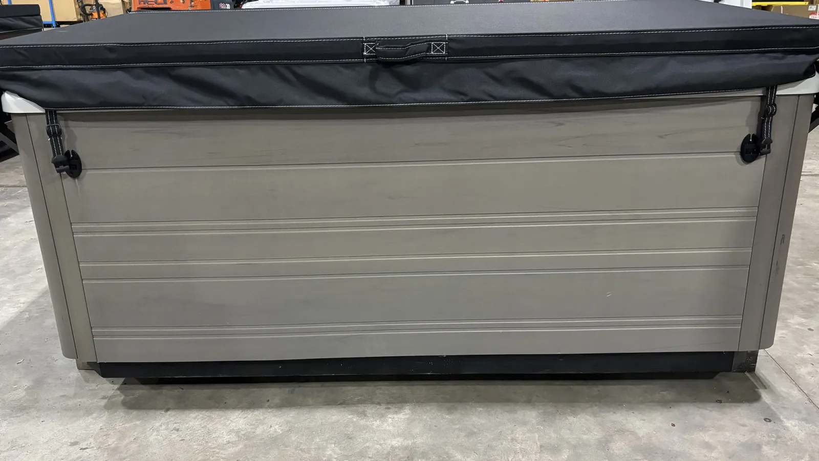 Rectangular gray hot tub with black cover secured by straps in industrial warehouse setting