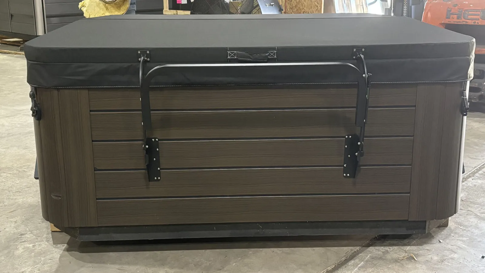 Closed brown wooden hot tub with black cover secured by metal brackets indoors on concrete floor