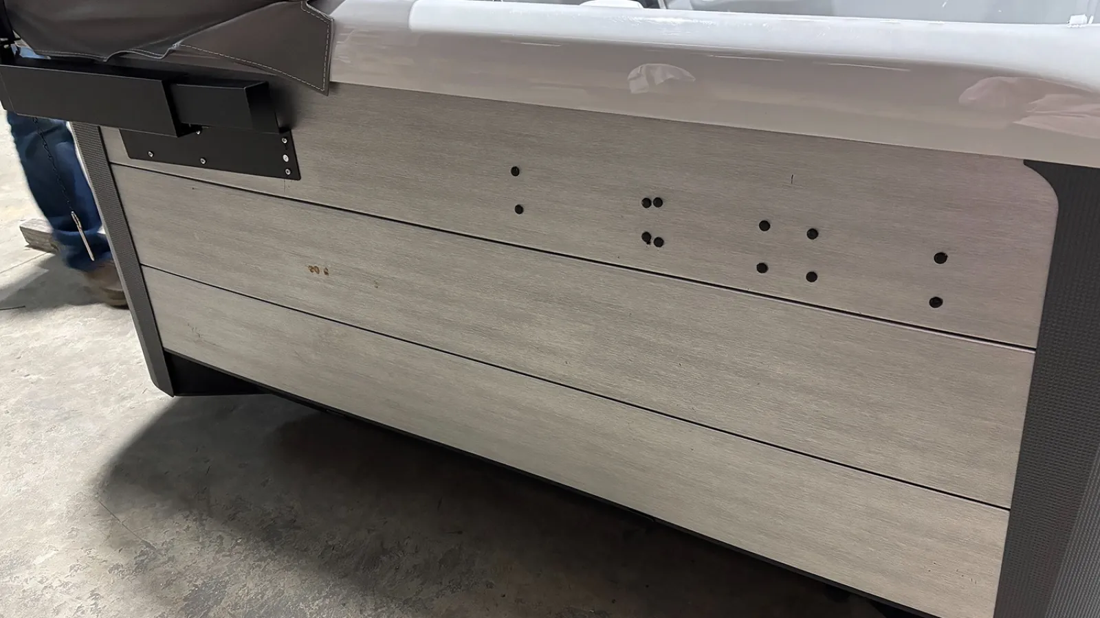 Side panel of a boat with light wood finish and mounting holes beneath gray cushioned seating on concrete floor.