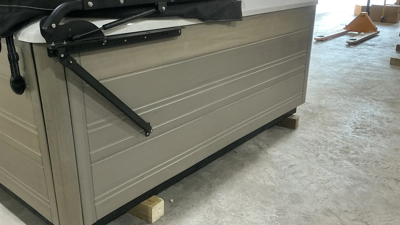 Side view of a closed hot tub with beige panels and a black cover hinge system indoors on concrete floor.