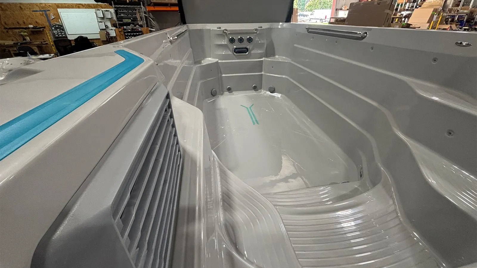 Empty modern white swim spa with seating, jets, and control panel in a warehouse setting under bright light.