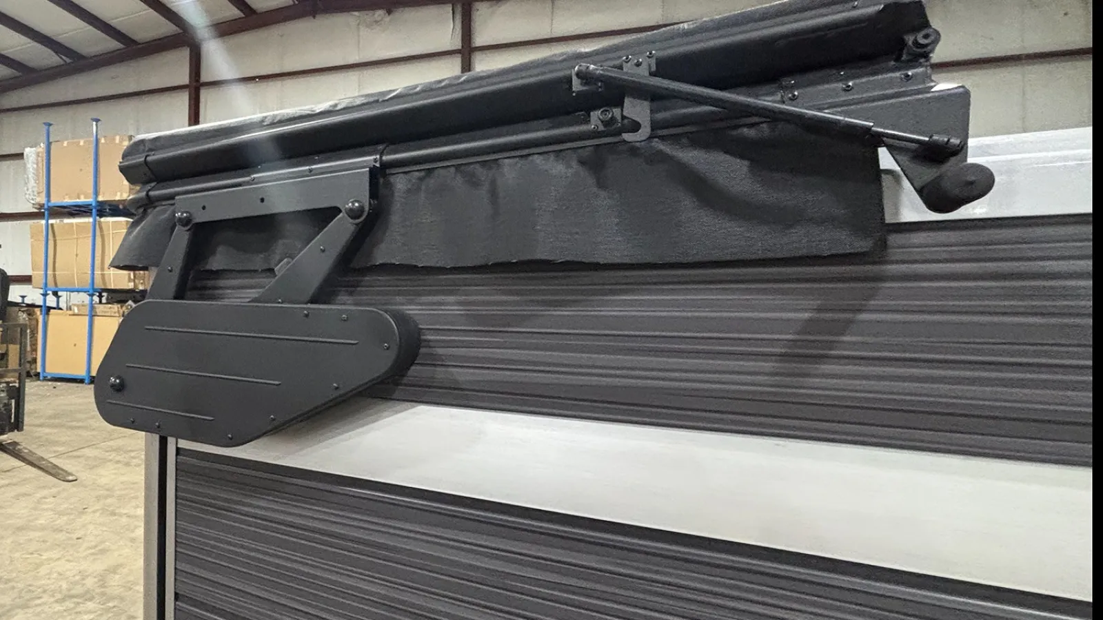 Black RV awning arm and rolled fabric attached to a dark gray ribbed vehicle side in a warehouse.