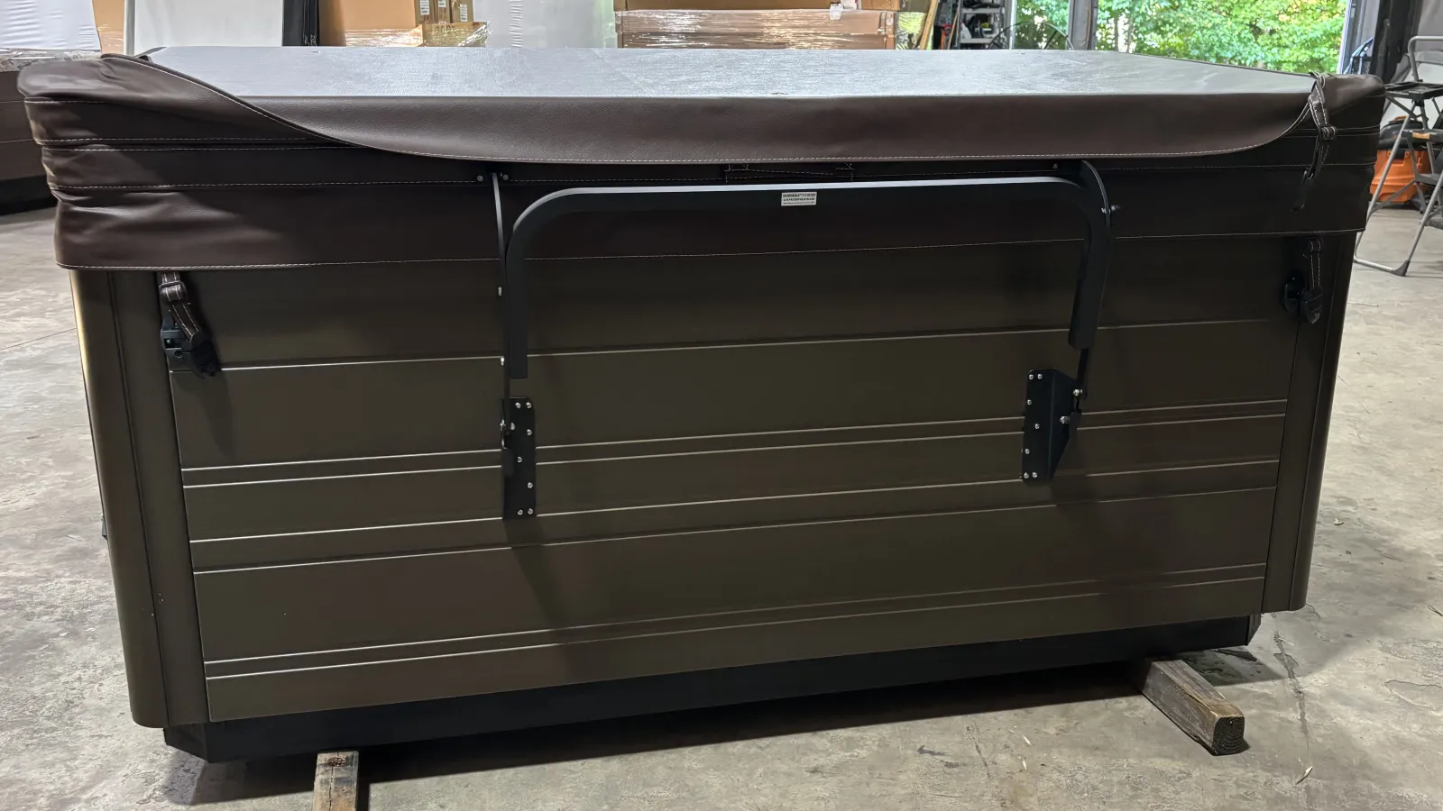 Side view of a brown hot tub with a closed cover and a metal handle, placed in an indoor storage area.