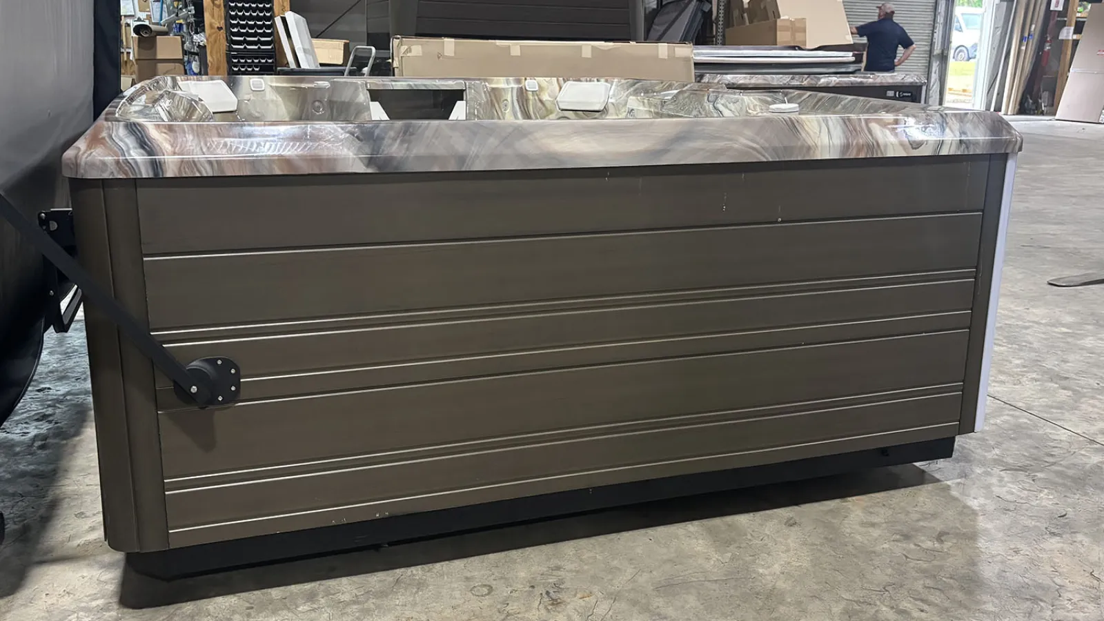 Modern brown and gray hot tub with marbled interior, set in a spacious warehouse with boxes and equipment.
