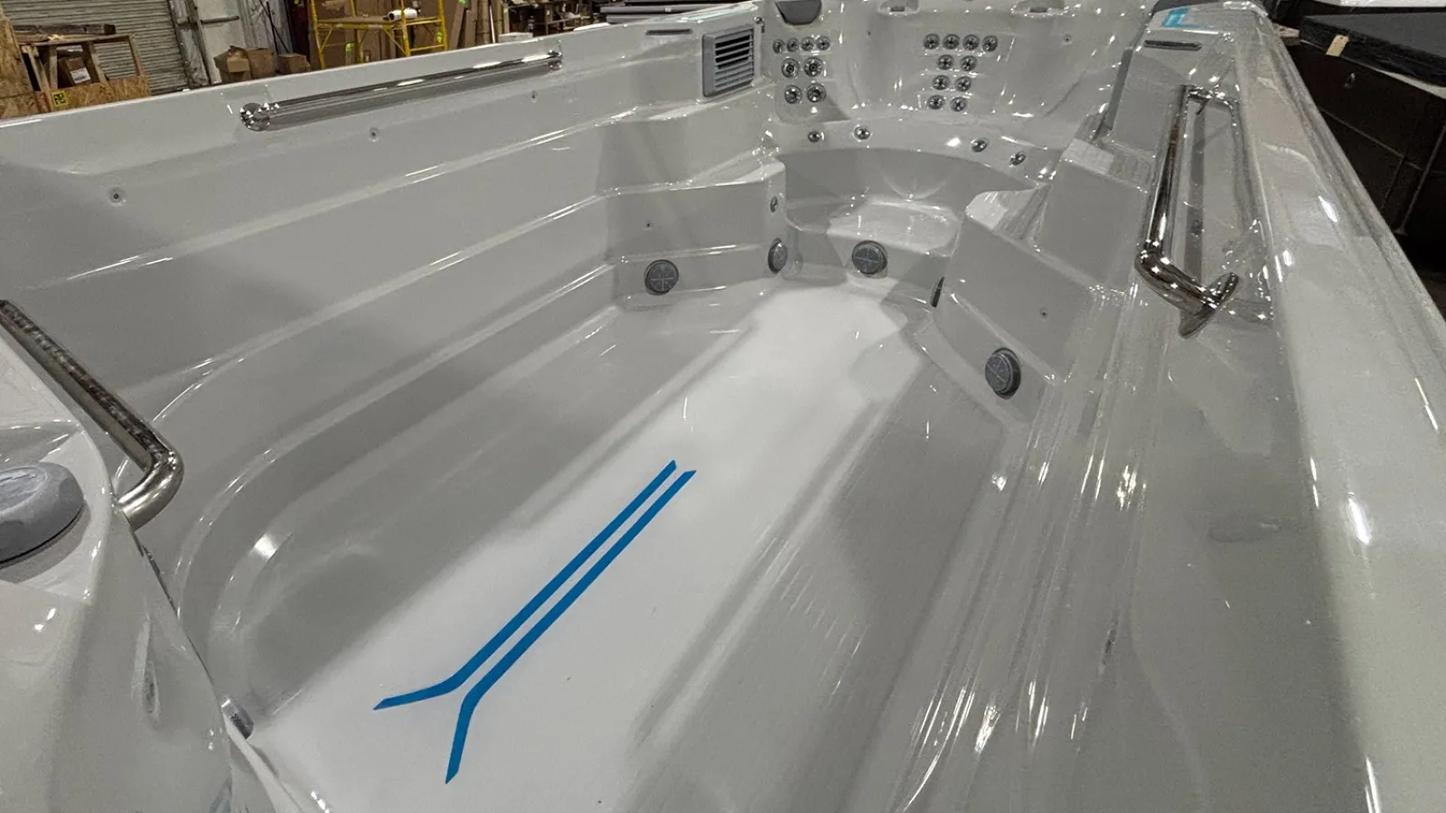 Close-up of a white swim spa with blue floor markings, multiple water jets, and handrails in an indoor warehouse.
