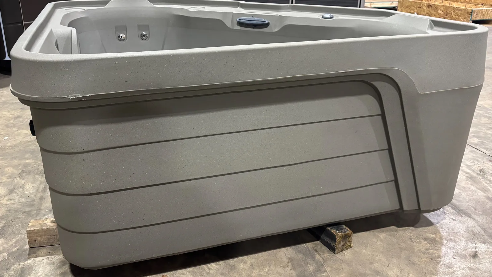 Gray rectangular outdoor hot tub with molded seats and jets on a concrete floor in a warehouse.