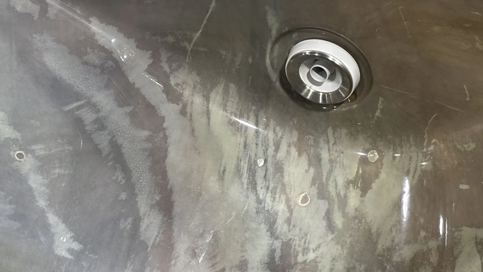 Close-up of a metal sink drain with scratched and stained basin surface under bright light.