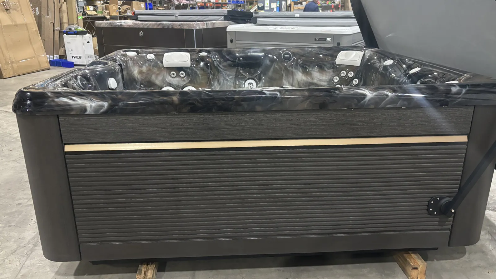 Modern black and gray hot tub with jets and headrests in a warehouse setting on wooden pallets