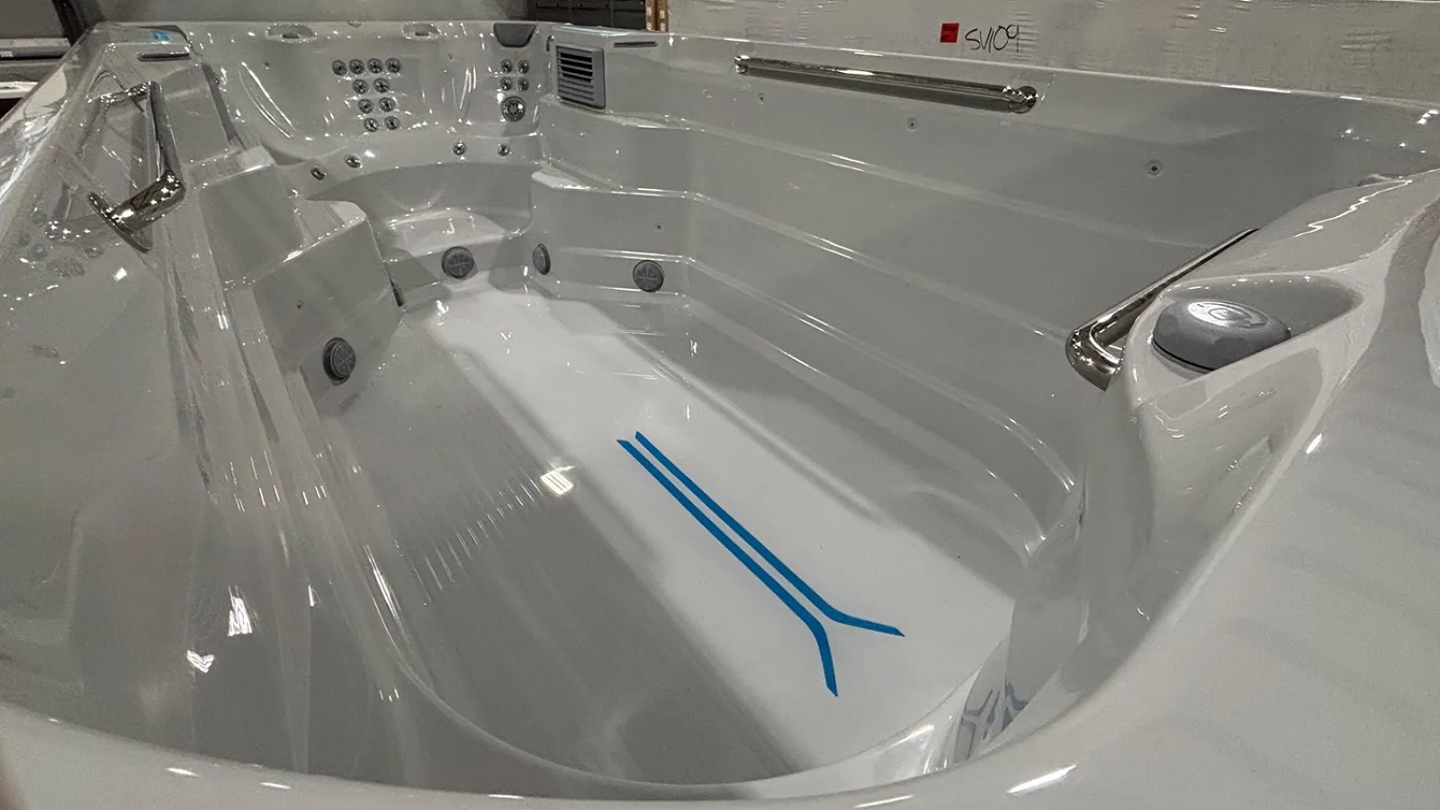 White modern swim spa with built-in seating, multiple water jets, handrails, and a blue lane marker at the bottom.