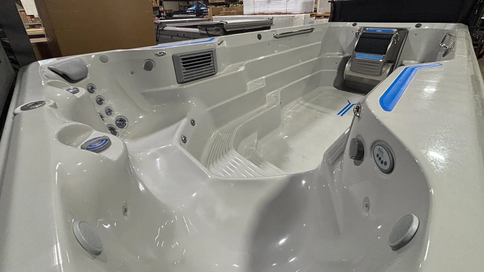 Large white swim spa with multiple seats, jets, and built-in controls in an indoor warehouse setting.