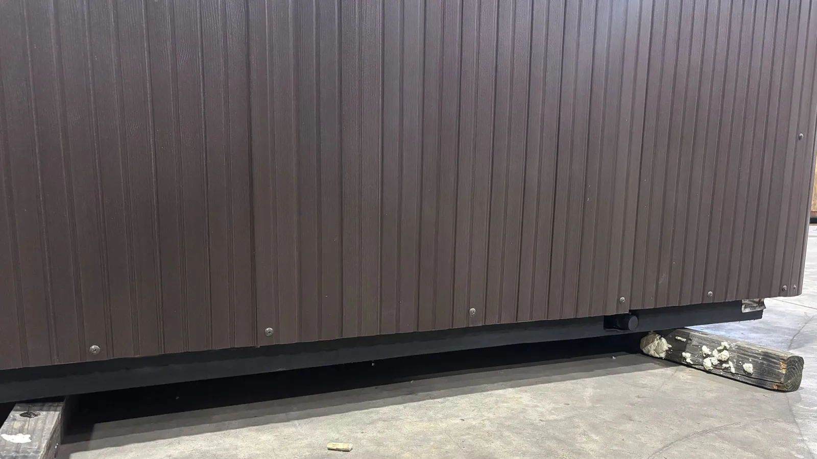 Close-up of the lower brown metal wall panel secured on wooden blocks on a concrete floor indoors.