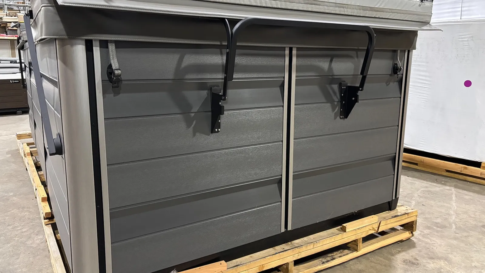 Gray outdoor storage box on wooden pallet in warehouse setting with metal support arms and textured panels.