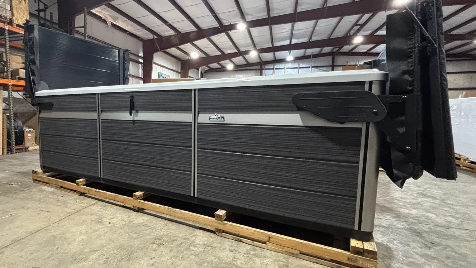 Large rectangular swim spa with dark gray panels and white top set on wooden pallet in warehouse.