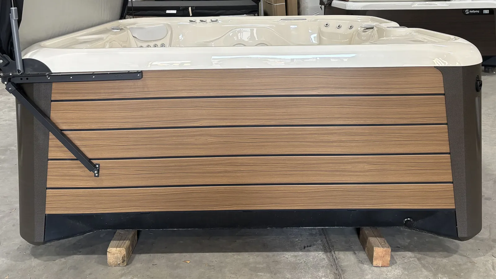 Modern outdoor hot tub with wood panel siding and beige interior jets on wooden blocks indoors.