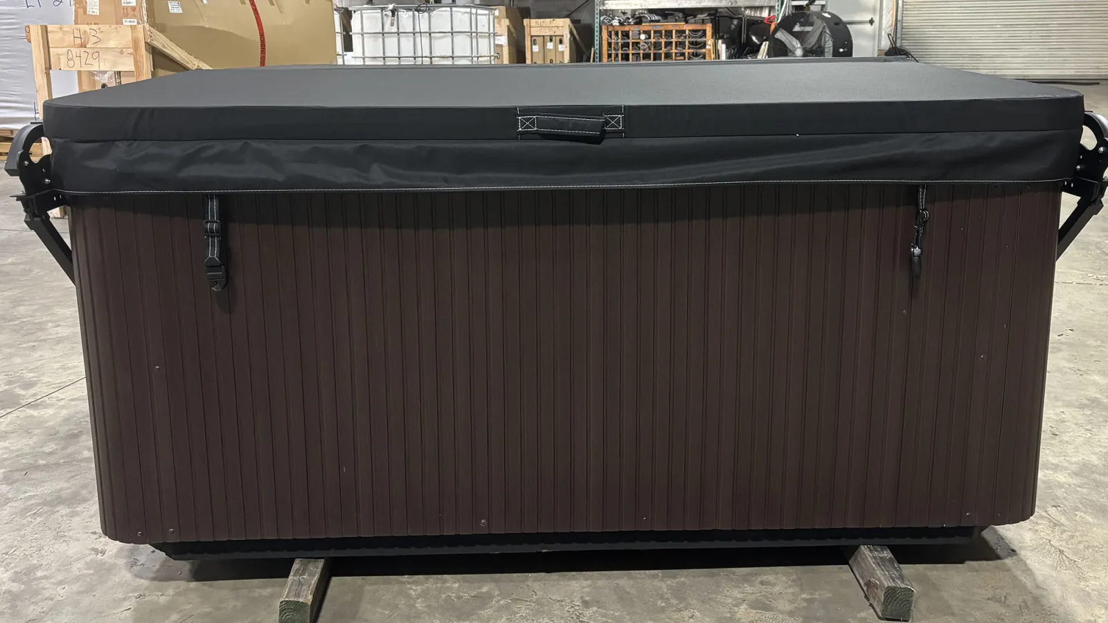 Closed brown hot tub with a black cover inside a warehouse surrounded by boxes and equipment.