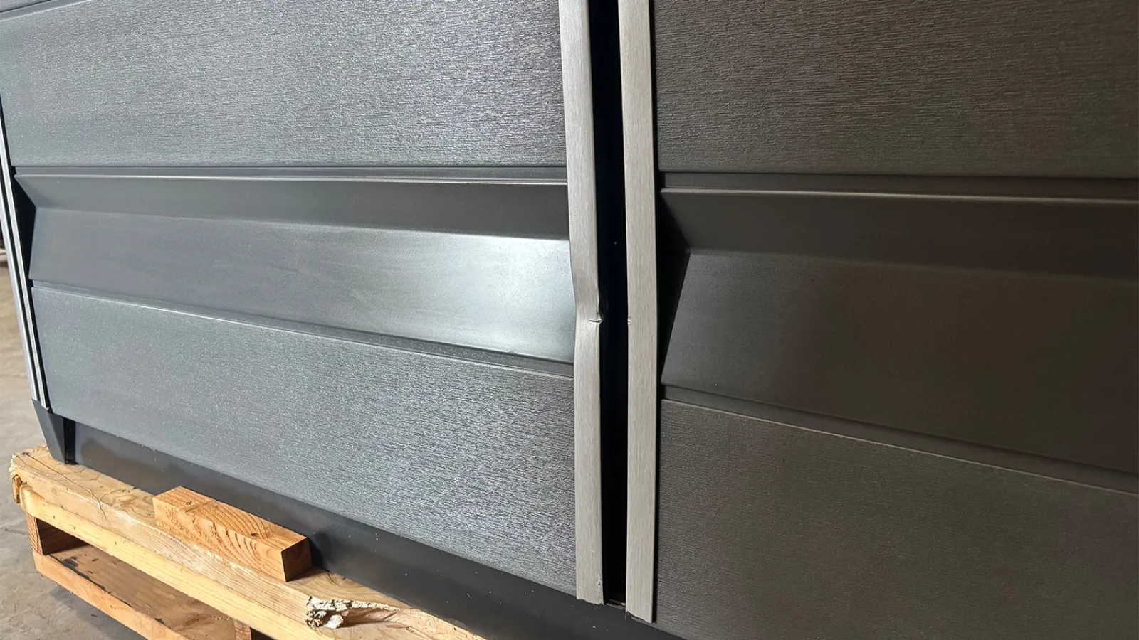 Close-up of black metallic panels with a slight gap, resting on a wooden pallet in industrial lighting.