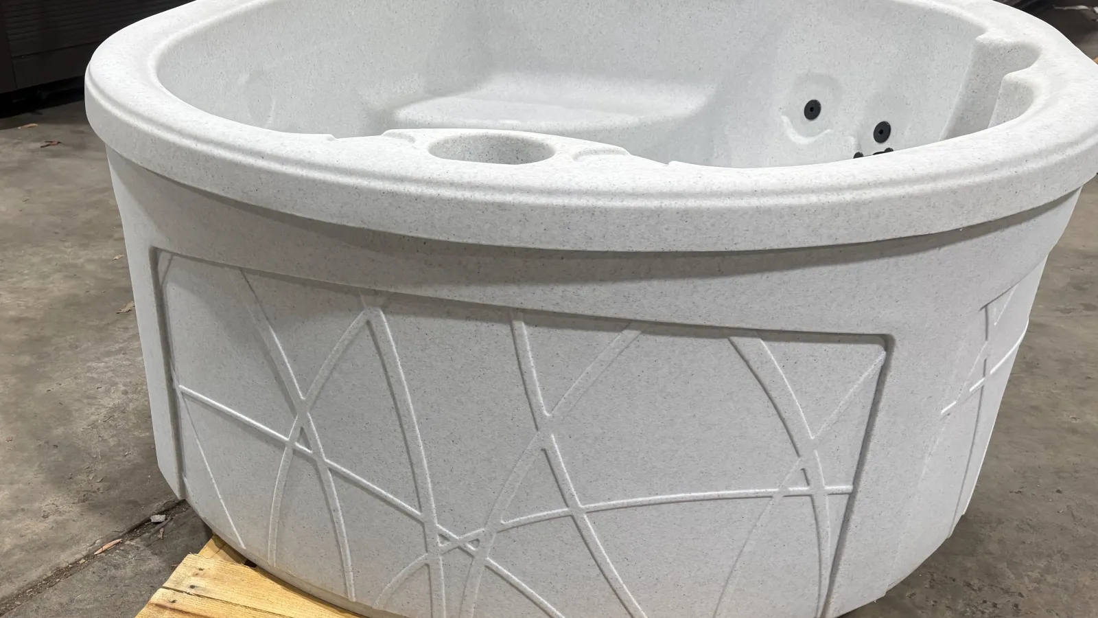 Round white hot tub with modern geometric patterns on exterior, standing on wooden pallet indoors.