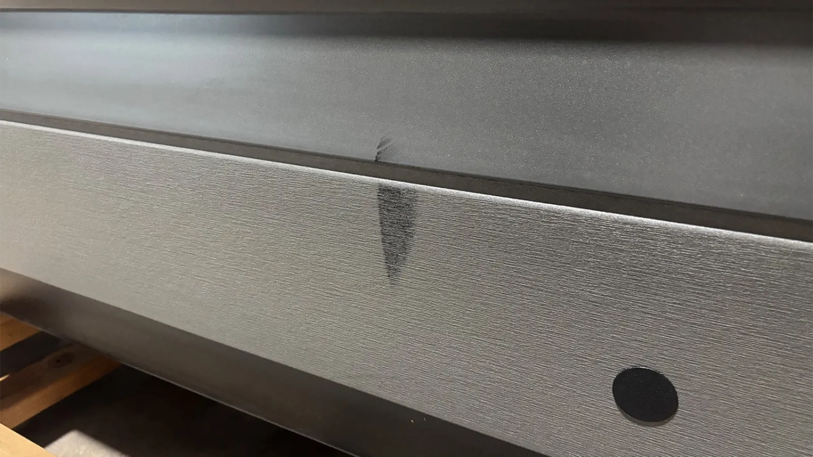 Close-up of metallic surface with a black spot and a smudge on textured stainless steel panel indoors.