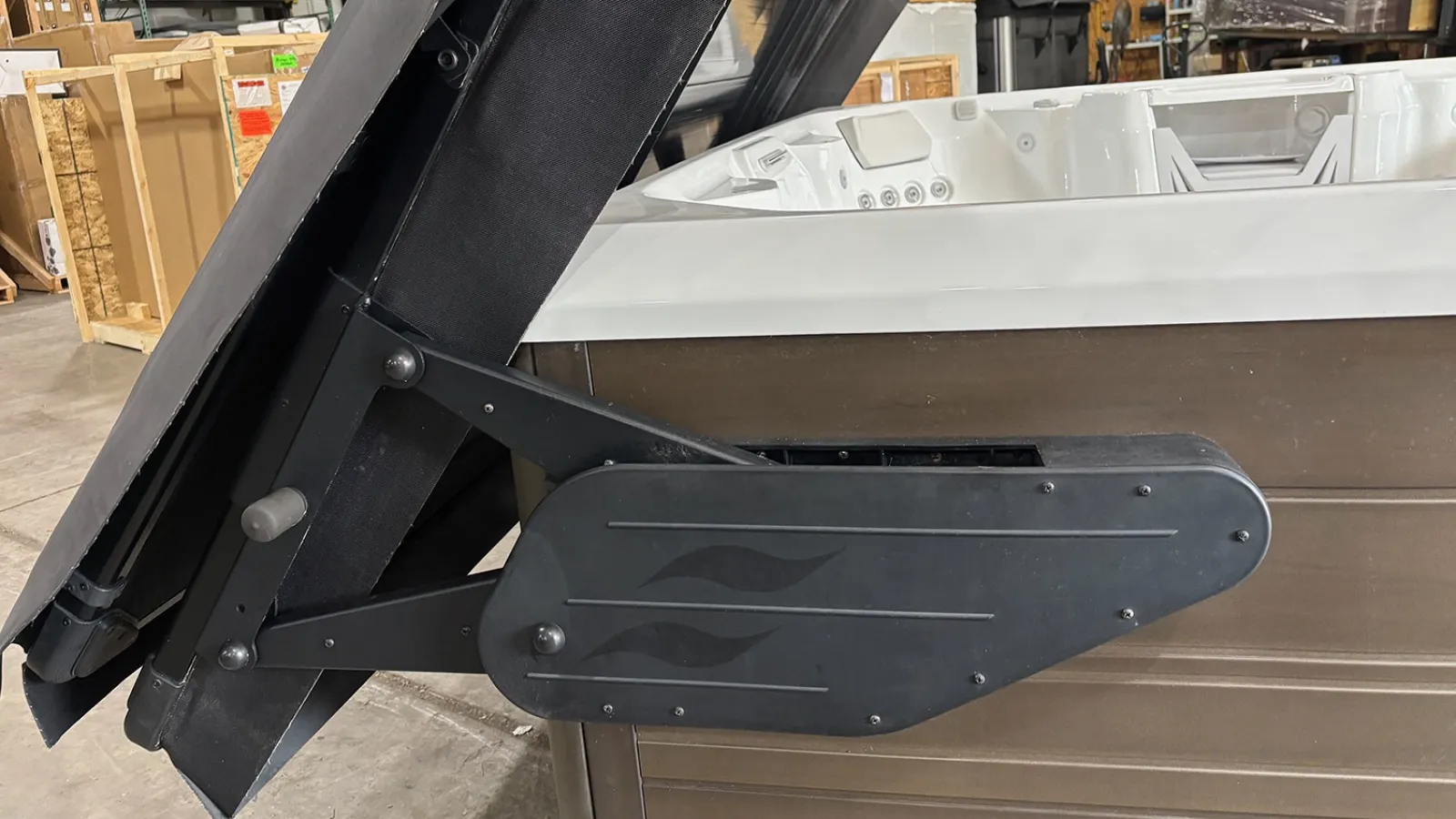 Black metal hinge mechanism attached to a brown hot tub cover in a warehouse setting with boxes in the background