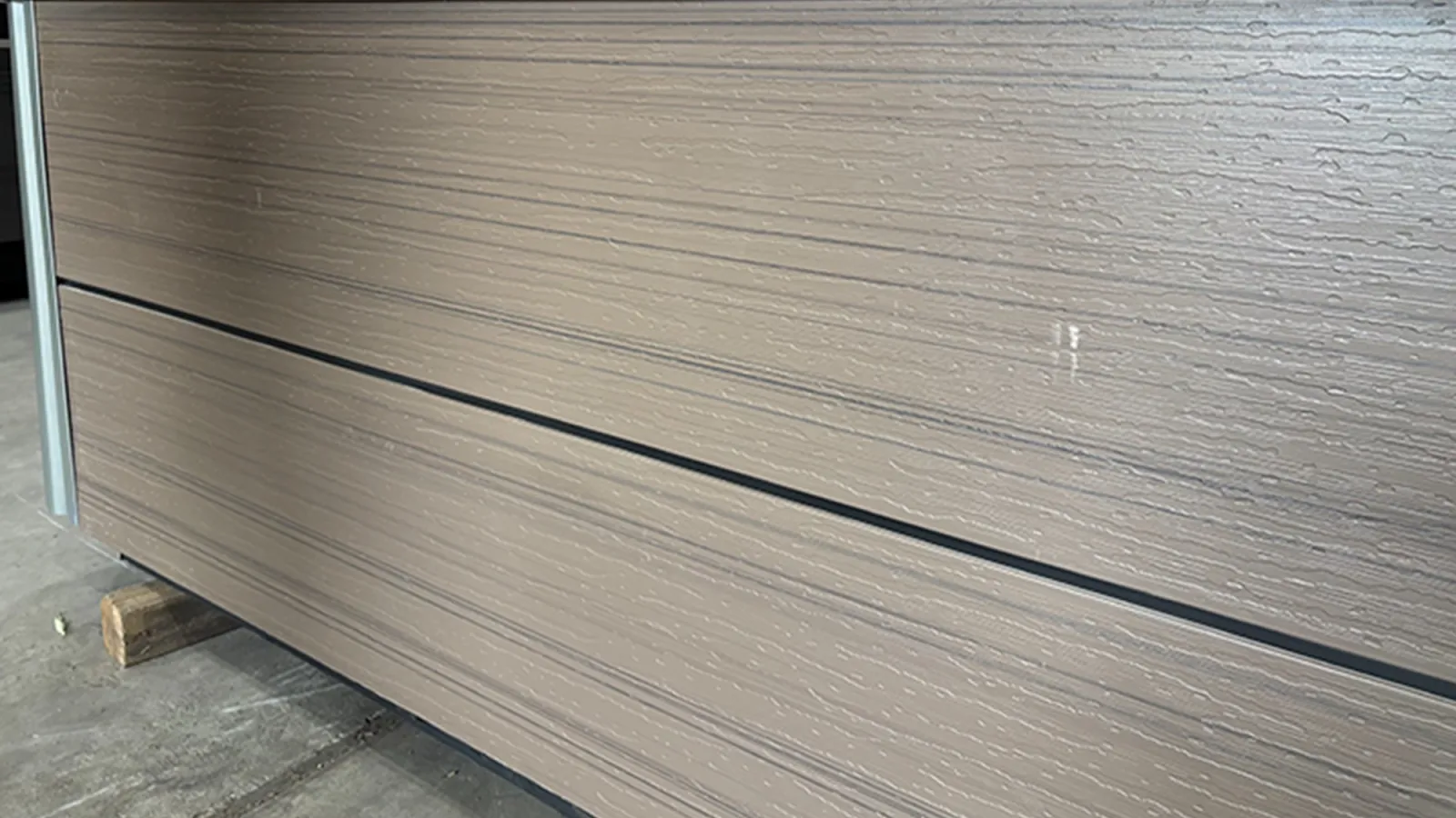 Close-up of textured beige horizontal siding panels with black trim lines on concrete floor indoors.