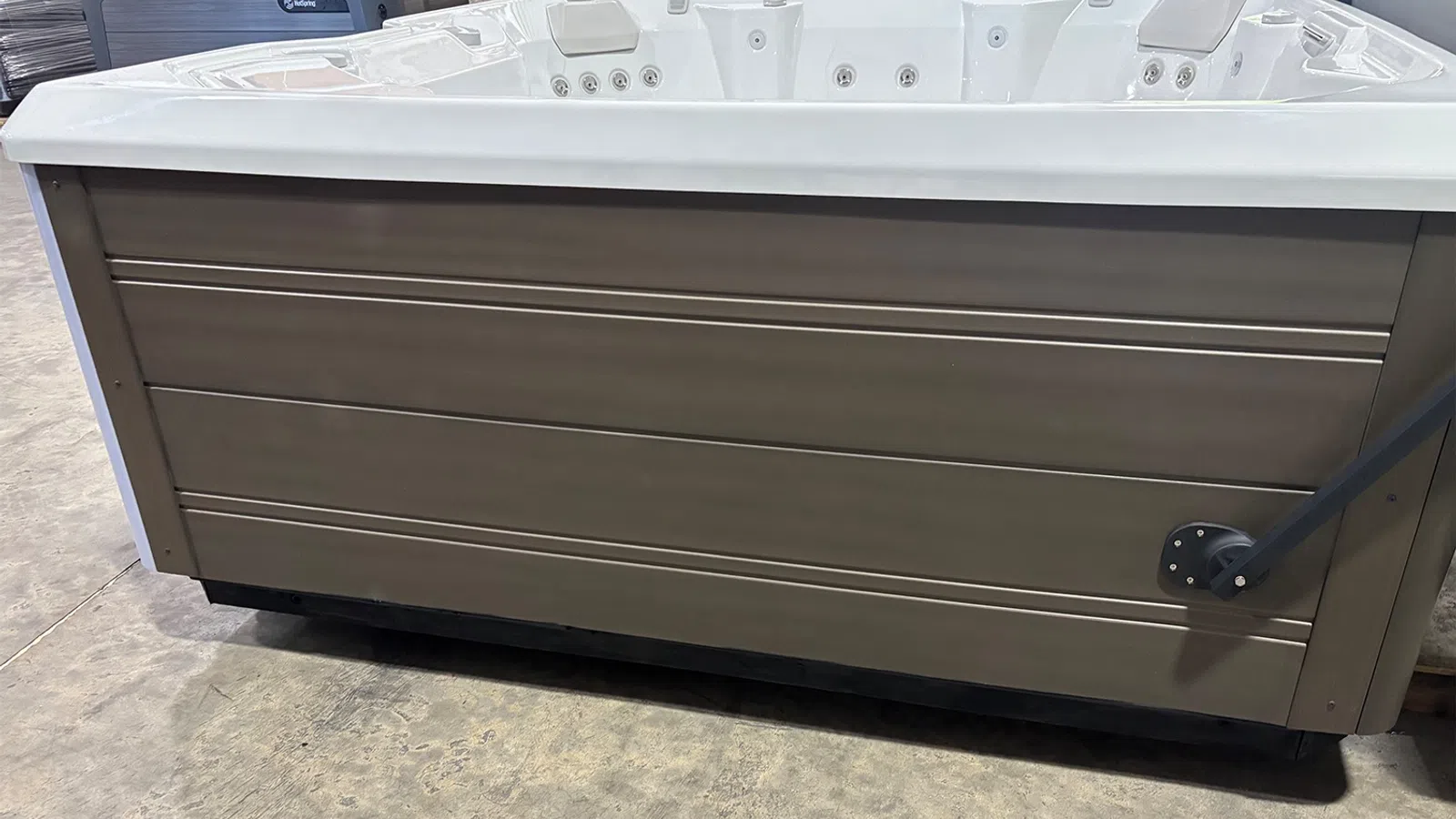 Large outdoor hot tub with gray horizontal paneling and multiple seating jets, placed on concrete floor.
