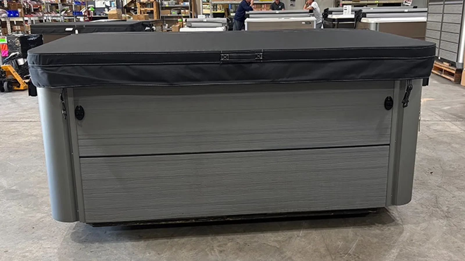 Rectangular gray hot tub with black cover inside a warehouse with workers and equipment.