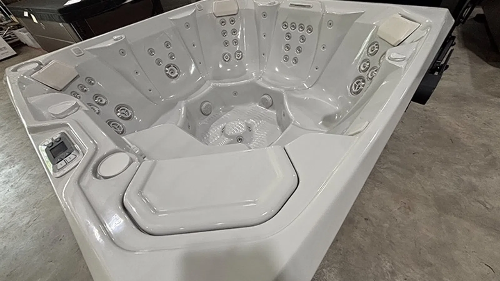 Modern white corner hot tub with multiple jet seats and digital control panel on a concrete floor.