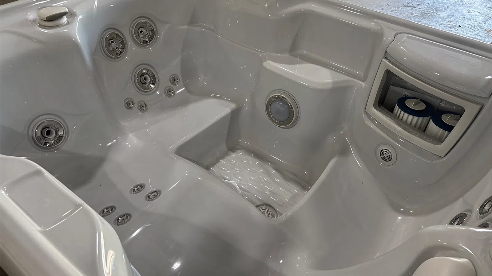 Empty white hot tub with multiple jets, built-in seating, and a filter compartment in a well-lit indoor space