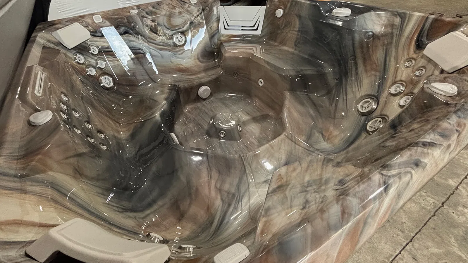 Marbled gray and beige empty hot tub with multiple jet nozzles and seats on a concrete floor indoors