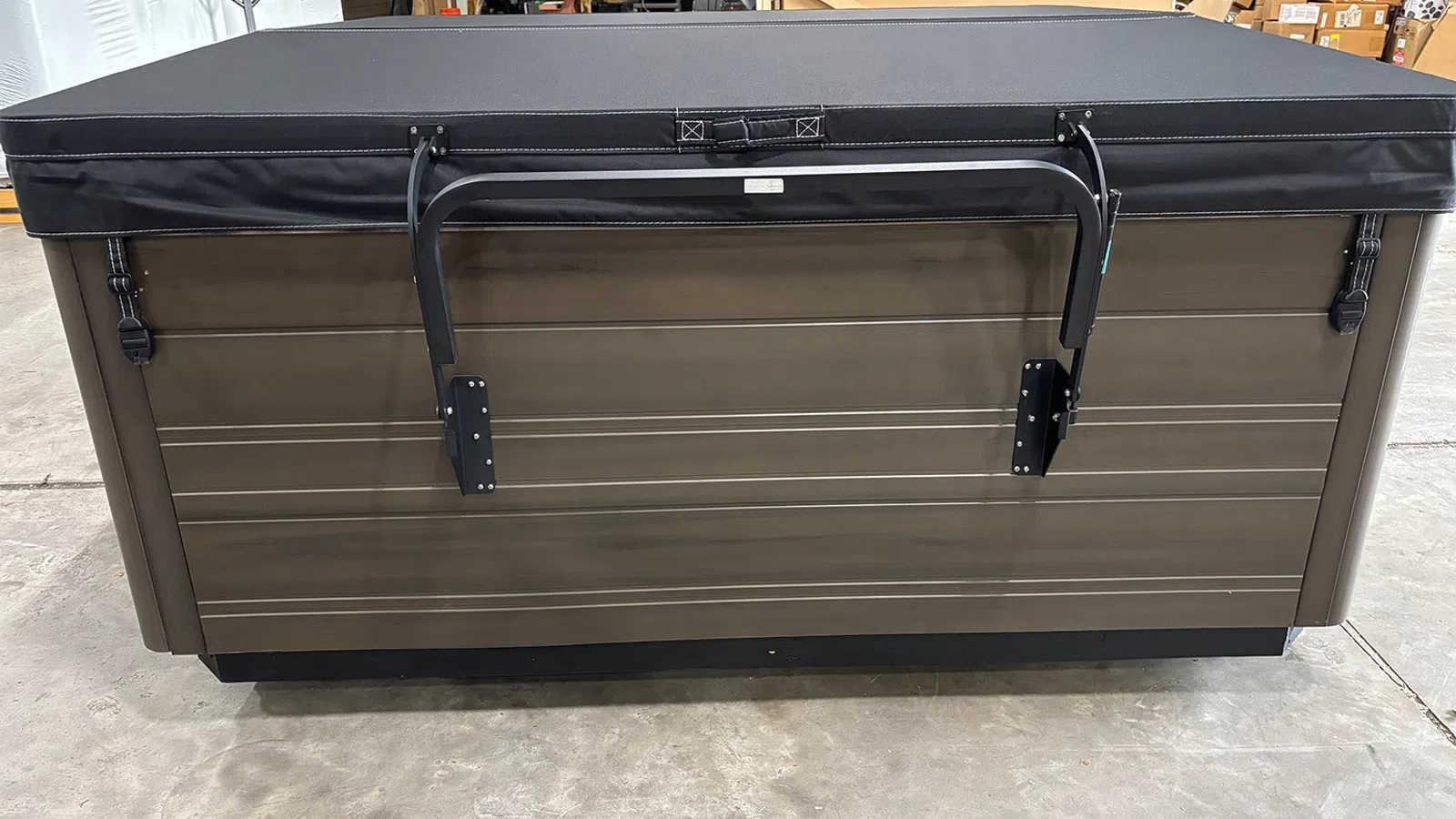 Brown hot tub with a closed black cover secured by two metal clamps inside a warehouse.