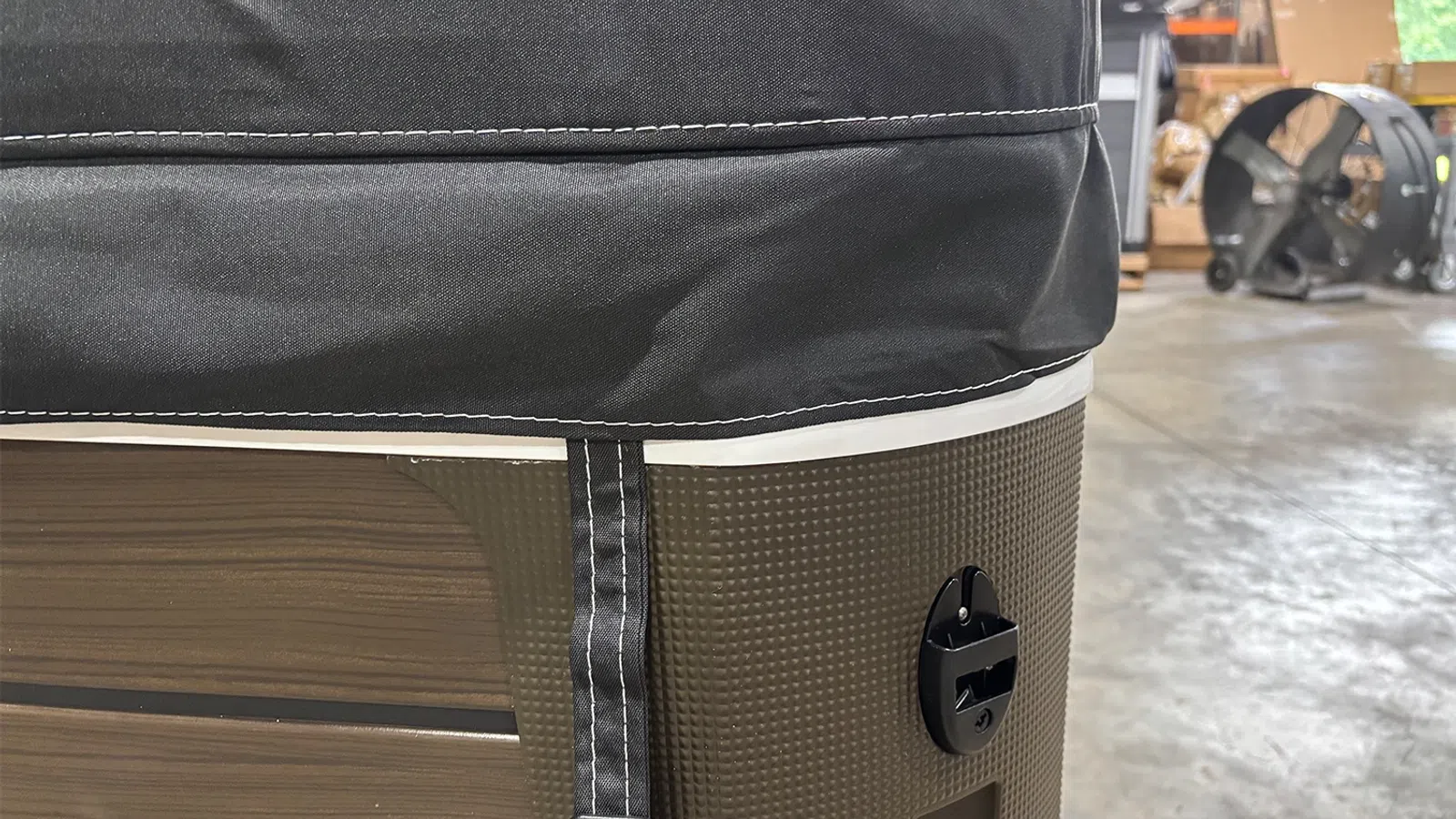 Close-up of a brown textured portable hot tub with black cover and strap inside a warehouse.