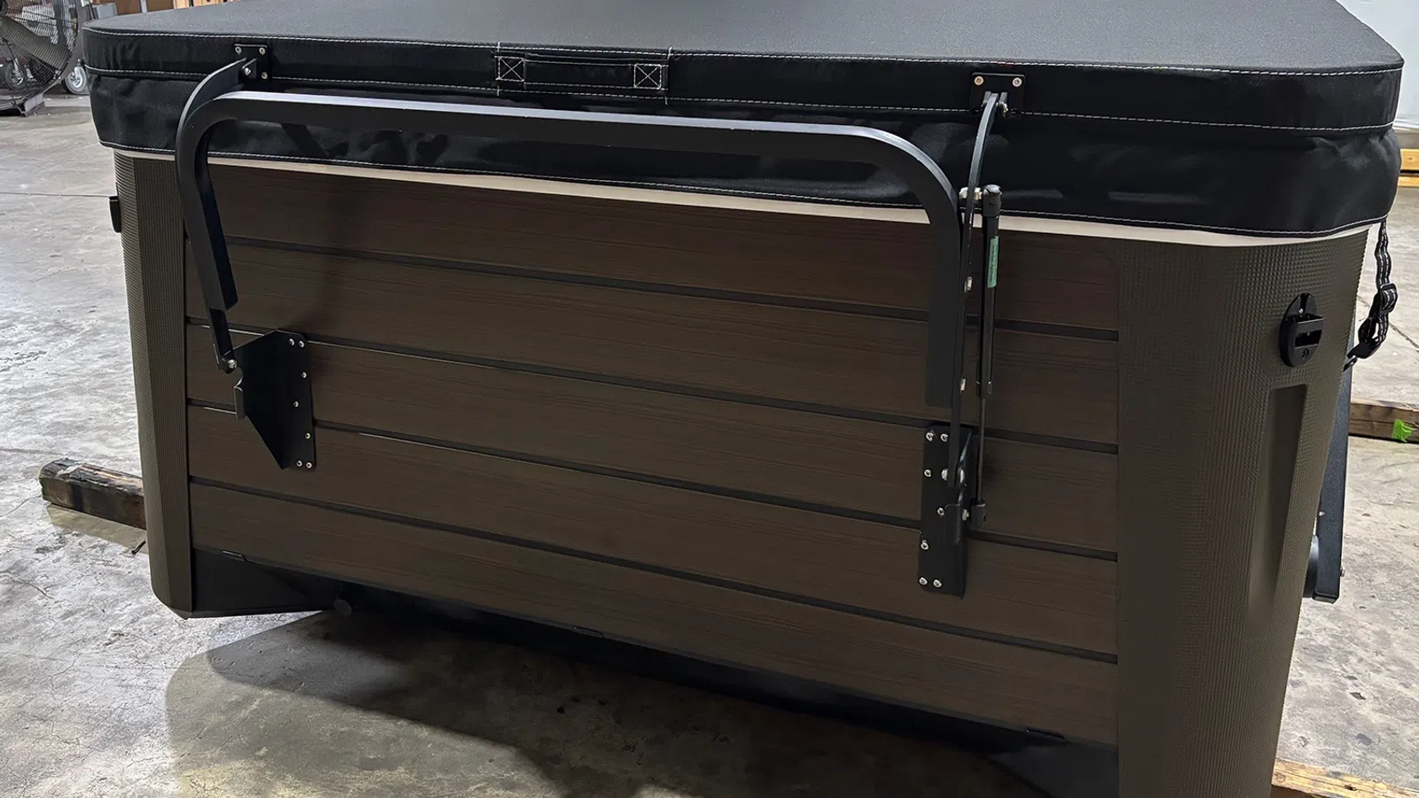 Side view of a brown hot tub with a black cover and metal lifting handle in a warehouse setting.