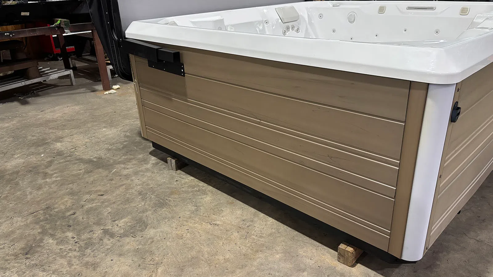 Close-up of a white and beige hot tub on wooden blocks in a garage workshop setting with tools in background.