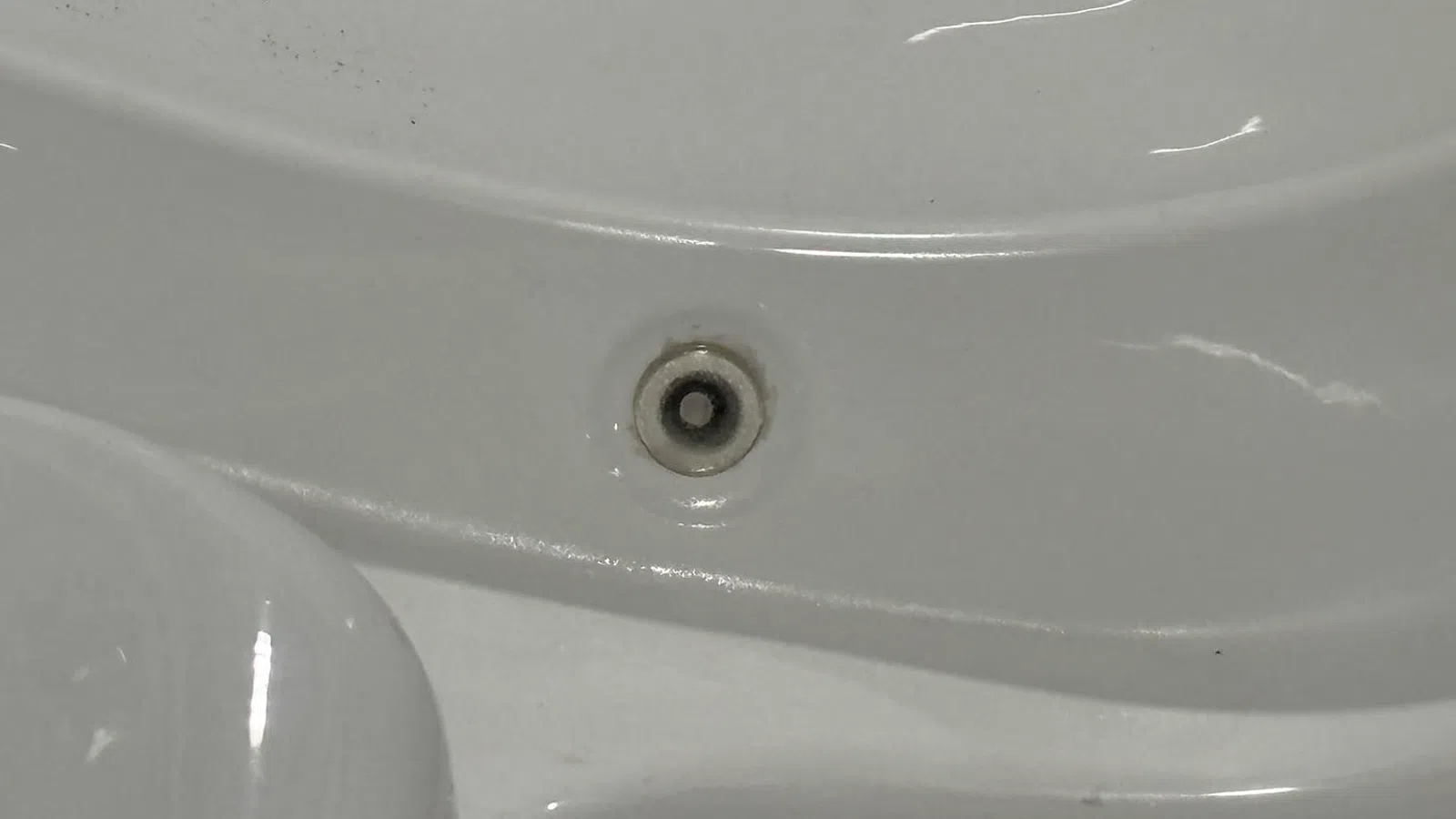 Close-up of a drain hole on a white glossy bathtub surface with highlighted reflections and contours.