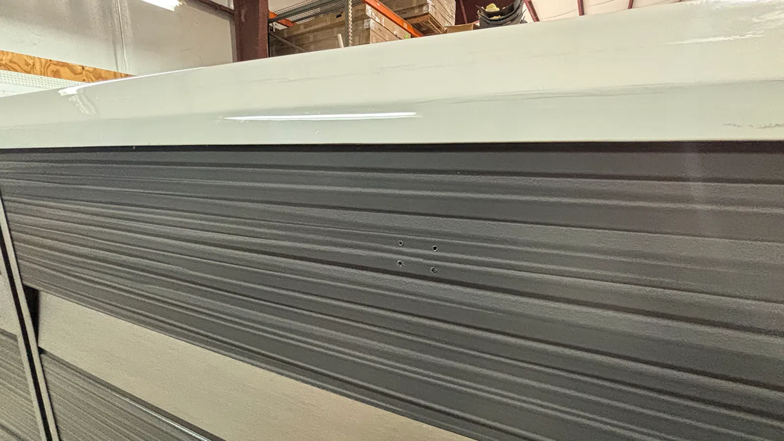 Close-up of a textured metal panel with horizontal ridges inside a warehouse with boxes above.