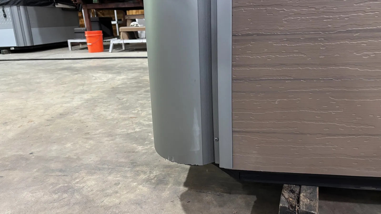 Close-up of the corner of a hot tub with textured wood-like paneling and a gray rounded edge on a cement floor.