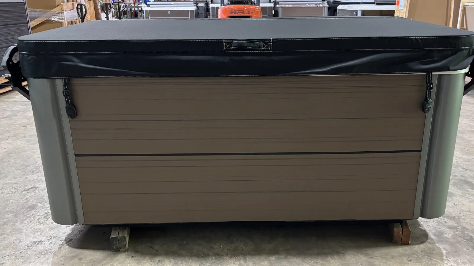 Closed outdoor hot tub with brown paneling and black cover on warehouse floor.