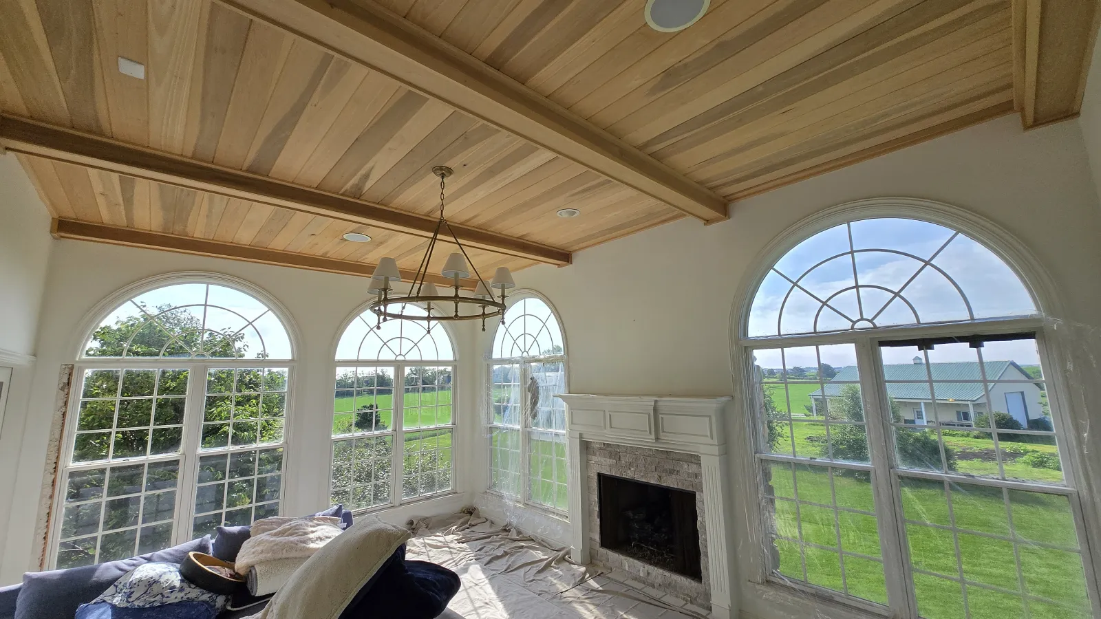 Bright sunroom with large arched windows, wooden ceiling, chandelier, and stone fireplace overlooking green fields.