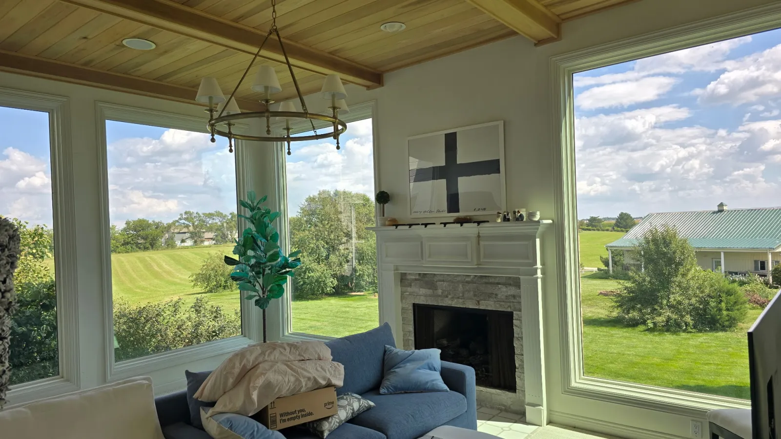 Cozy living room with large windows overlooking green fields, a fireplace, blue sofa, and wooden ceiling.