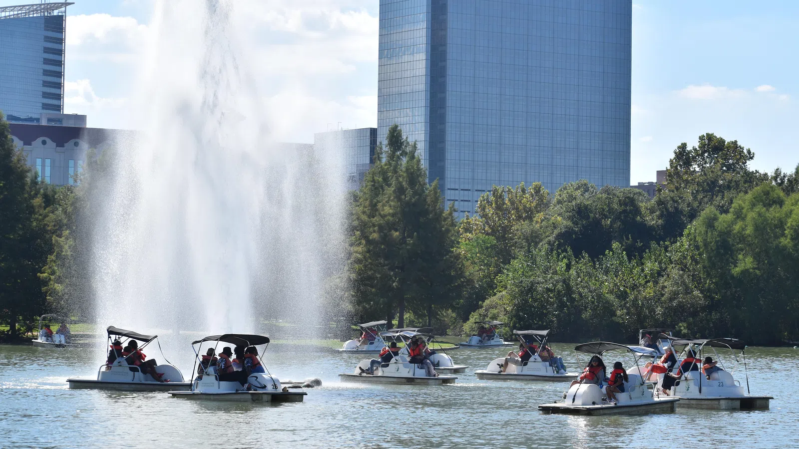 Multiple pedal boats with people wearing life jackets on a lake near a large fountain and urban skyscrapers on a sunny day