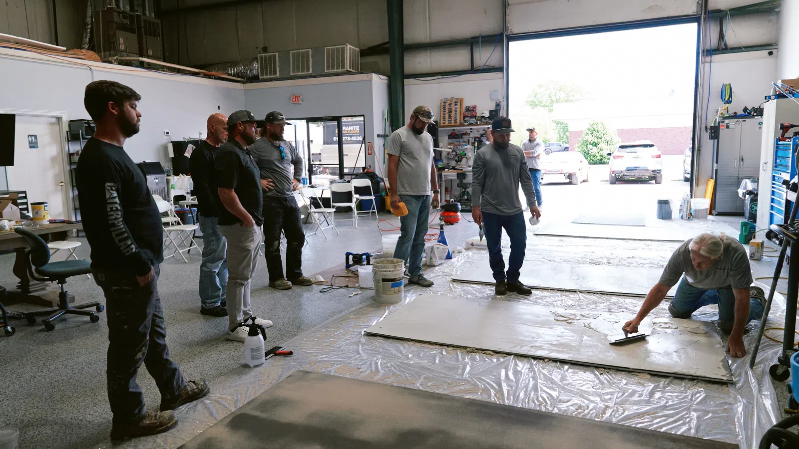 Group of men observing a worker smoothing large slabs on the floor inside a workshop with open garage door.