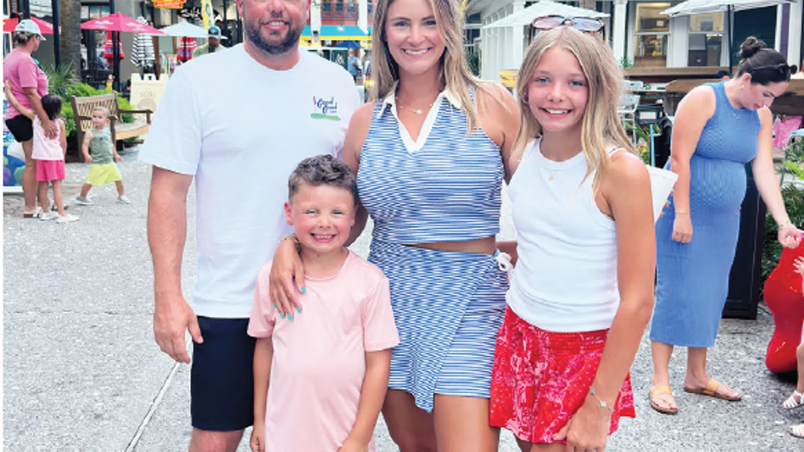 Smiling family of four posing under red and white umbrellas on a sunny shopping street with palm trees.