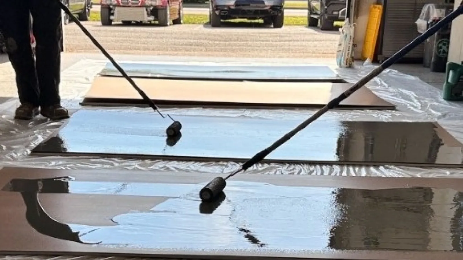 Two workers use rollers to apply black epoxy coating on wooden panels in a garage with cars outside.