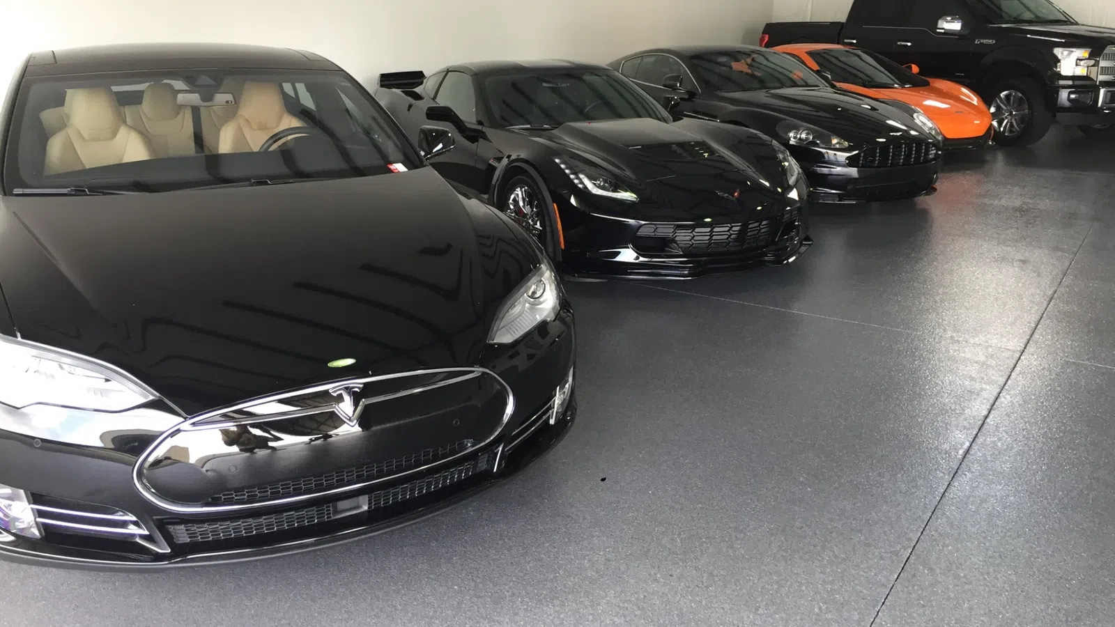 Row of luxury and sports cars parked indoors including black Tesla, Corvette, and an orange sports car.