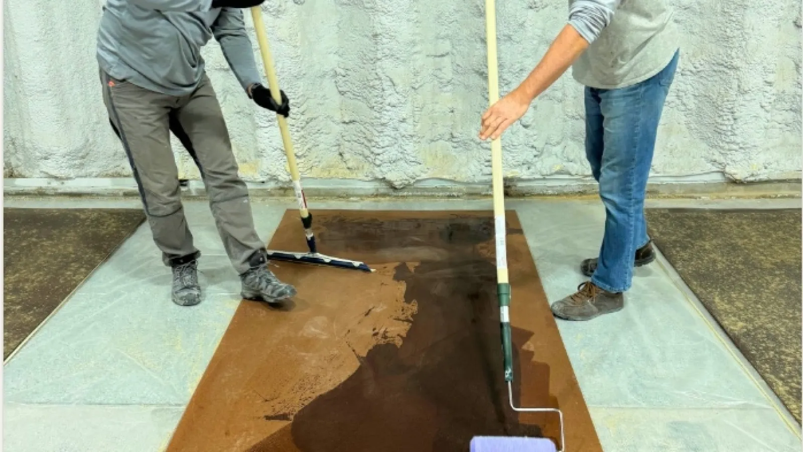 Two men using rollers and brushes to apply dark stain on a large wooden panel in an industrial setting.