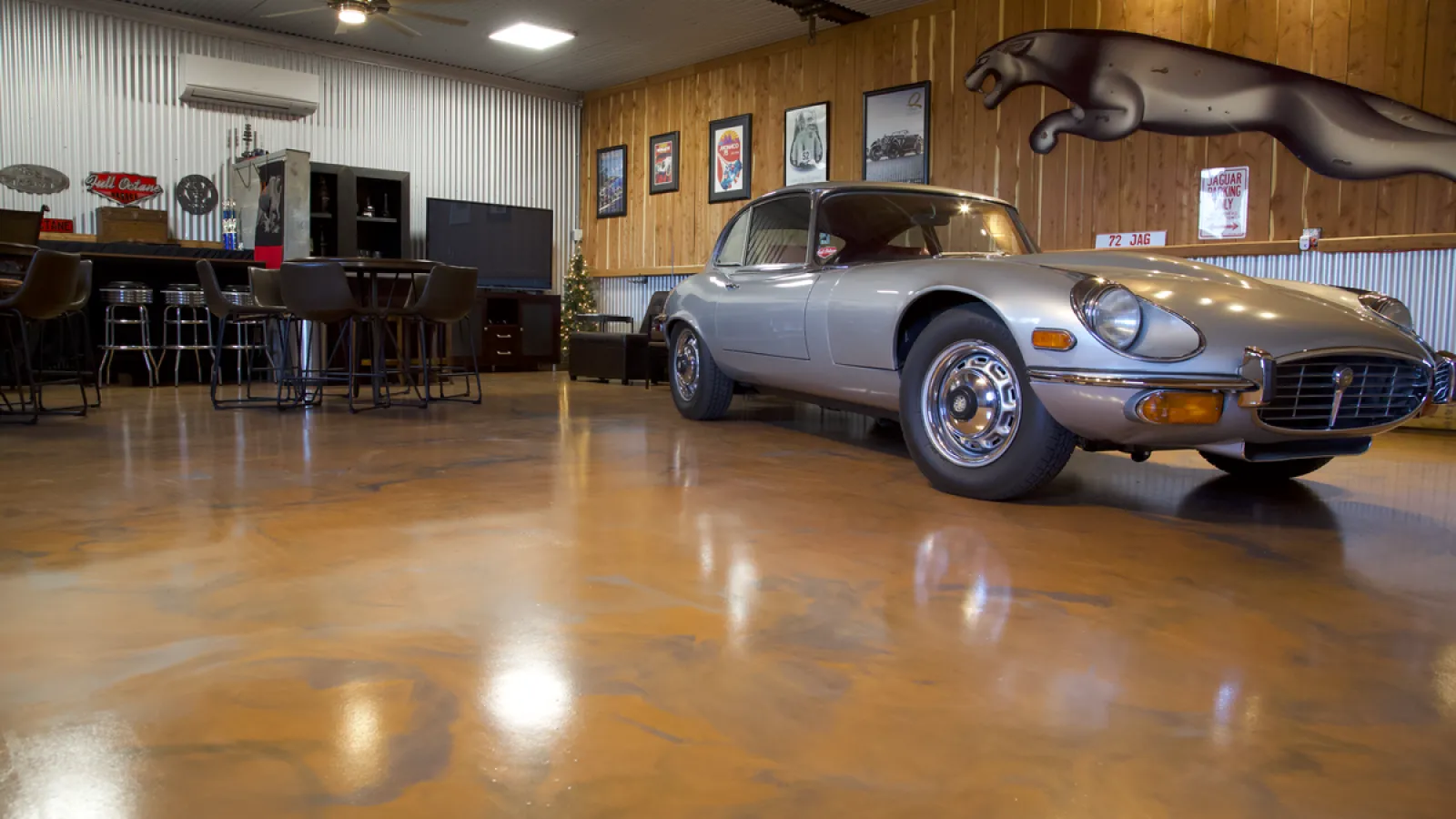 Classic silver Jaguar E-Type coupe parked on polished floor in stylish automotive-themed garage with bar and vintage decor