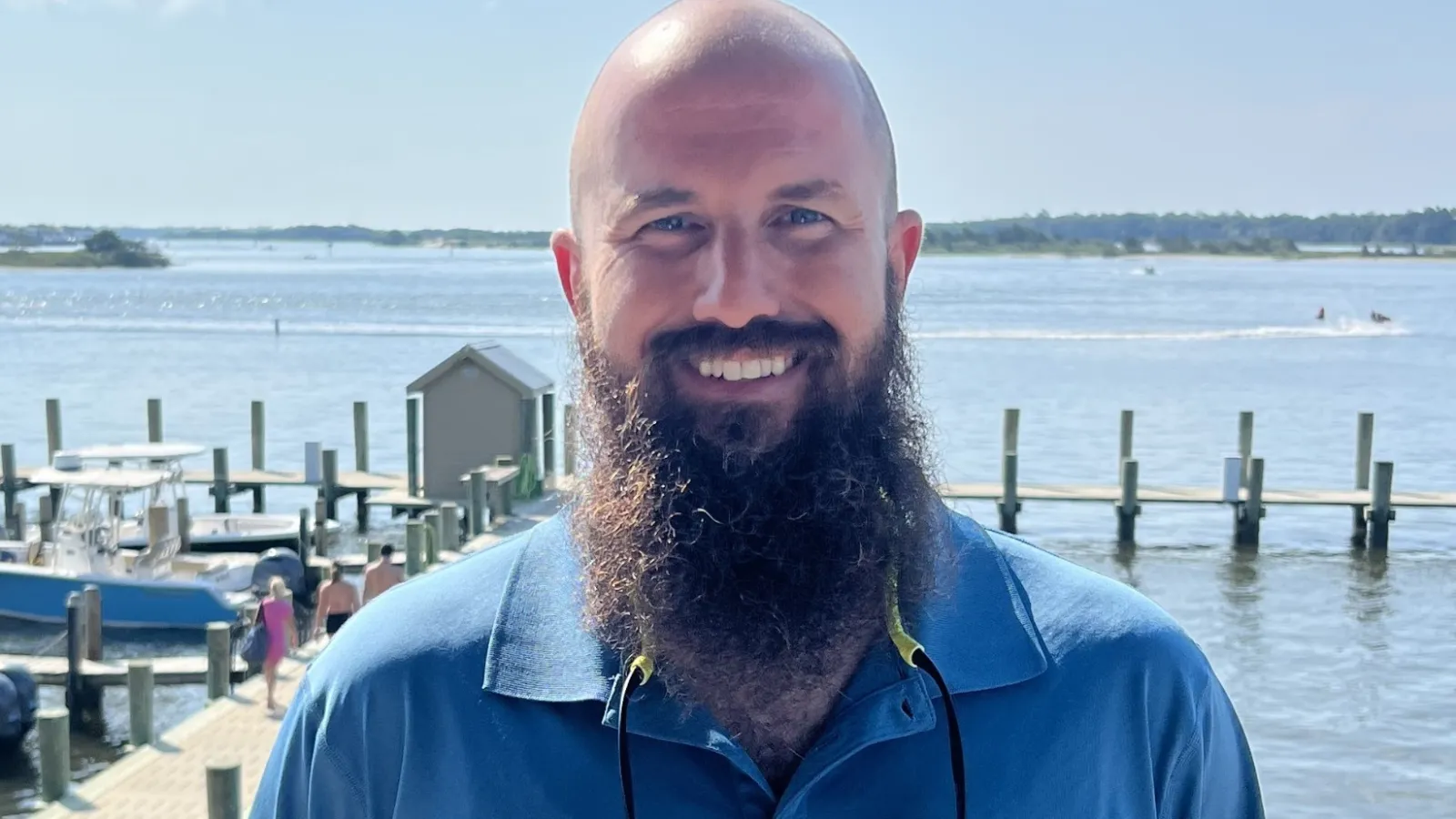 Smiling man with a long beard wearing a blue Columbia shirt standing by a sunny waterfront with docks and boats.