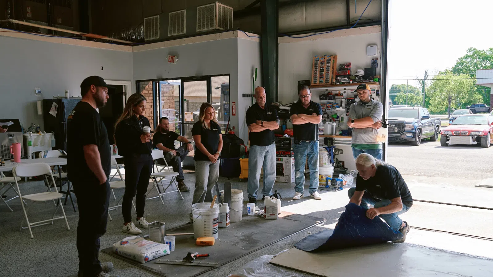 Group of people watching a man demonstrating a flooring or coating technique inside a workshop garage.
